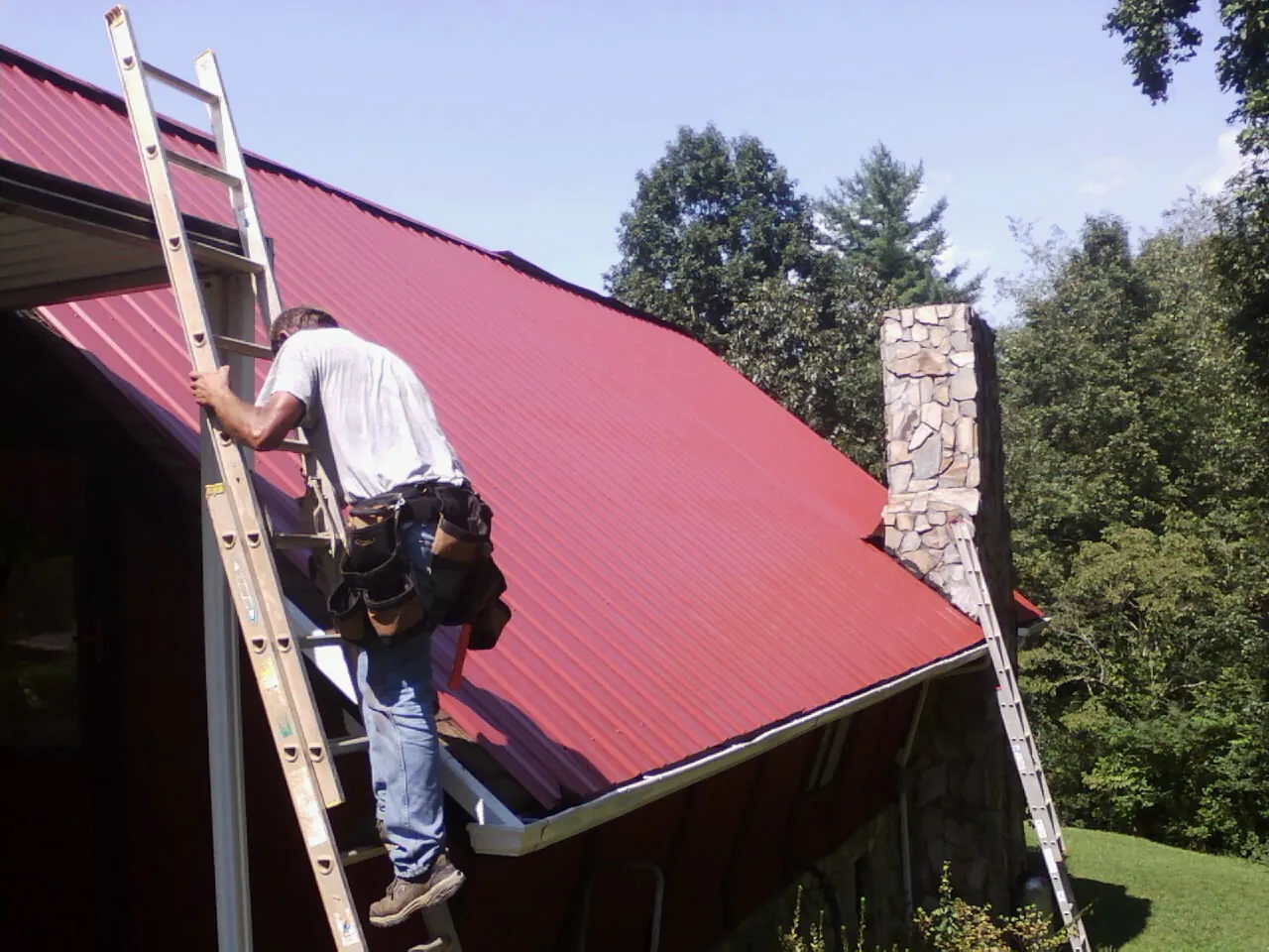 Roofer on a ladder beside a red metal roof and stone chimney — Soffit & Fascia Repair by Roofing Brothers in Hollidaysburg